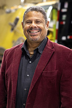 Portrait of Eric Dorcean. He is smiling at the camera and wearing a dark gray button-down shirt under a burgundy jacket.
