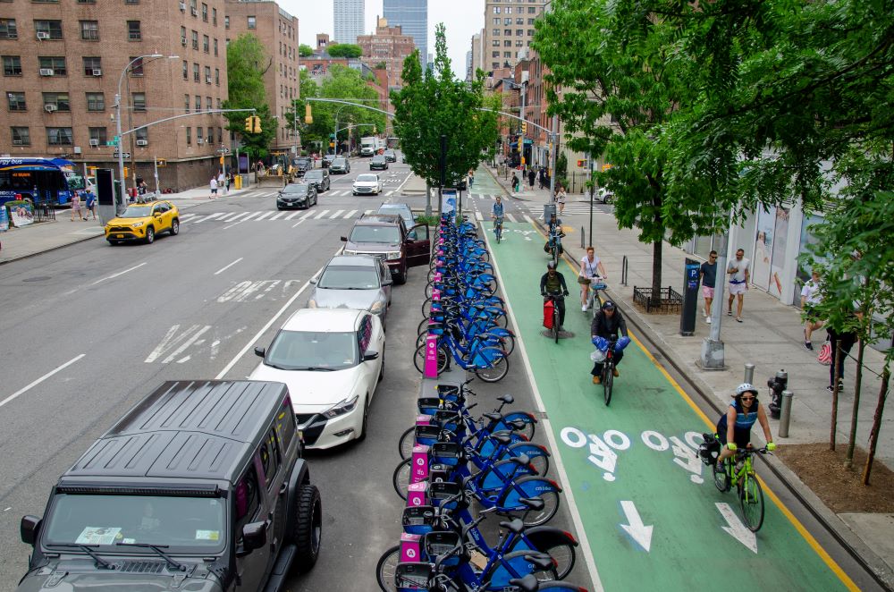 Multiple cyclists ride on a wide green bike lane on Ninth Avenue in Manhattan.
